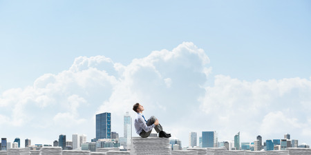 Thoughtful businessman sitting on pile of documents and looking away with cityscape on background. Mixed media.の写真素材