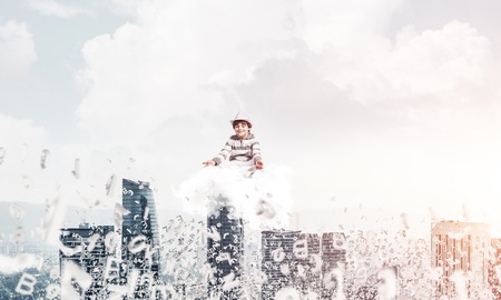 Young little boy keeping eyes closed and looking concentrated while meditating on cloud among flying letters with cityscape view on background.の写真素材