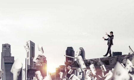 Businessman walking blindfolded among flying documents on concrete bridge with huge gap as symbol of hidden threats and risks. Cityscape and sunlight on background. 3D rendering.の写真素材