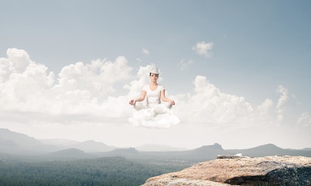 Young woman keeping eyes closed and looking concentrated while meditating on cloud in the air with beautiful and breathtaking landscape on background.の写真素材