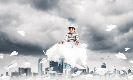 Young little boy keeping eyes closed and looking concentrated while meditating on cloud among flying paper planes with cityscape view on background.の写真素材