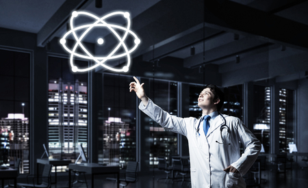 Conceptual image of successful and young doctor in white uniform interracting with molecular structure while standing against night cityscape view on background.の写真素材