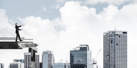 Businessman walking blindfolded on concrete bridge with huge gap as symbol of hidden threats and risks. Cityscape view on background. 3D rendering.の写真素材