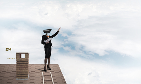 Faceless businesswoman with camera zoom instead of head standing on house roofの写真素材