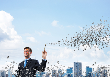 Happy and young businessman in black suit holding paintbrush in his hand and smiling while standing against modern cityscape view on backgroundの写真素材