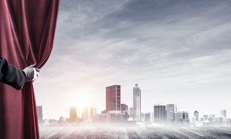 Human hand opens red velvet curtain to modern cityscape and cloudy skyの写真素材