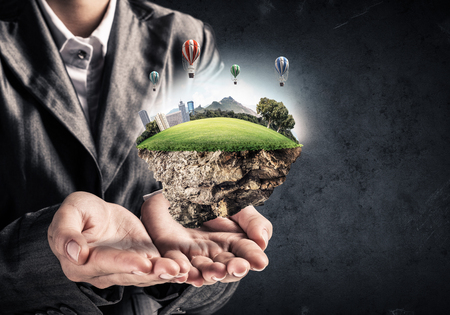 Businessman in suit keeping green island with skycraper city with flying balloons in hands and with gray wall on background.の写真素材