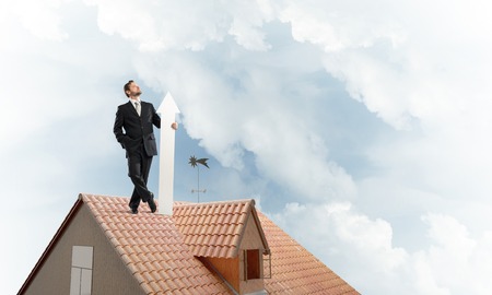 Conceptual image of young businessman in suit looking upside and holding big white arrow in hand while standing on brick roof with cloudy skyscape view on background.の写真素材