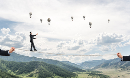 Businessman walking blindfolded on rope above high mountains as symbol of hidden threats and support. Flying balloons and nature view on background.の写真素材
