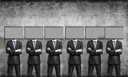 Businessmen in suits with TV instead of their heads keeping arms crossed while standing in a row in empty room with gray wall on background.の写真素材