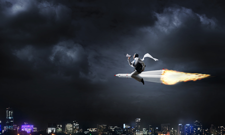 Conceptual image of young businessman in suit flying on rocket with night cityscape and blue sky on background.の写真素材