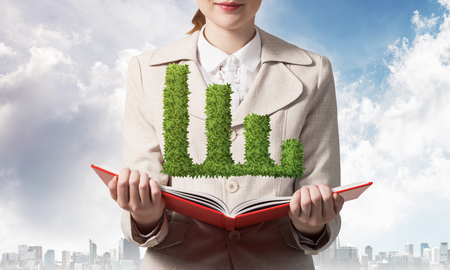 Businesswoman with green plant shaped financial graph above opened notebook. Business paperwork and accounting. Elegant young woman in white business suit on background of cityscape and blue sky.の写真素材
