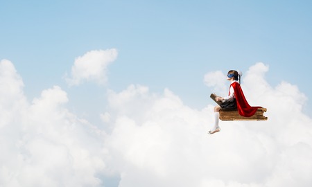 Little child in mask and cape floating on book in sky and readingの写真素材