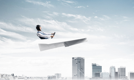 Businessman in aviator hat and goggles driving paper plane above downtown with high buildings. Modern megalopolis panorama with blue skyscape. Mixed media business concept. Dreaming and imagination.の写真素材