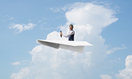 Pilot in leather helmet and goggles driving paper plane on background of blue cloudy sky. Traveling around the world. Funny man flying in small paper airplane. Sky panorama with fluffy clouds.の写真素材
