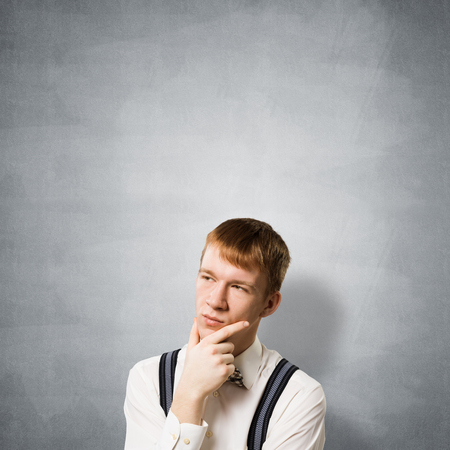 Redhead student looks pensively upwards and tries to remember something. Puzzled boy has serious facial expression. Portrait of guy wears shirt, bow tie and suspenders on background of grey wall.の写真素材