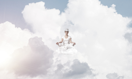 Woman in white clothing keeping eyes closed and looking concentrated while meditating on clouds in the air with cloudy skyscape on background.の写真素材