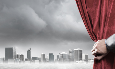 Human hand opens red velvet curtain to modern cityscape and cloudy skyの写真素材