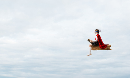 Little child in mask and cape floating on book in sky and readingの写真素材