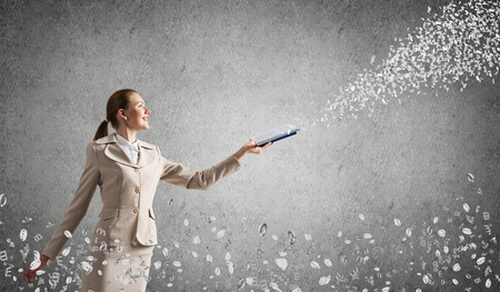 Woman looking at flying out random letters from opened book. Teaching and education. Information flow concept. Elegant young woman in white business suit with open notebook on background of grey wall.の写真素材