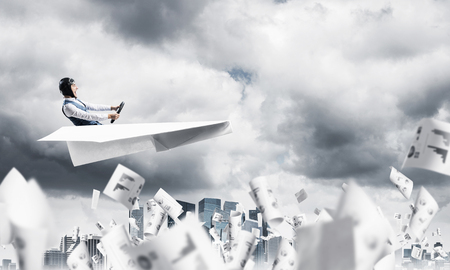 Business accounting and statistics concept with pilot. Aviator driving paper plane above falling paper sheets with infographics. Man flying in paper airplane above paperwork in dramatic cloudy sky.の写真素材