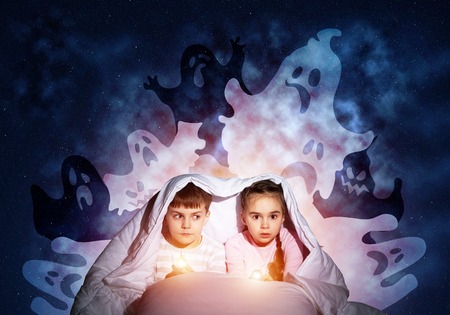 Scared girl and boy with flashlights hiding under blanket from imaginary phantoms. Frightened kids sitting in bed on night sky background. Children in pajamas and boo ghosts silhouettes above themの写真素材