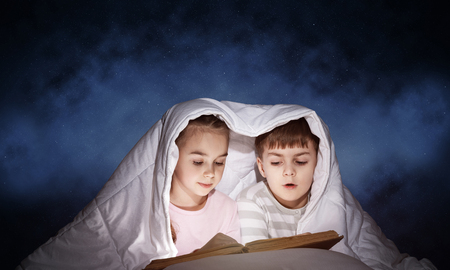 Engrossed little girl and boy reading book in bed after bedtime. Pretty kids together hiding under blanket. Preschool children in pajamas reading fairy tales. Bedroom with deep blue sky backgroundの写真素材
