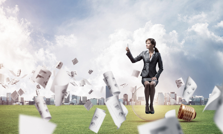 Young woman using smartphone on green field with falling paper documents. Pretty girl sitting on big glowing lamp. Businesswoman chatting with phone. Business statistics and analytics.の写真素材