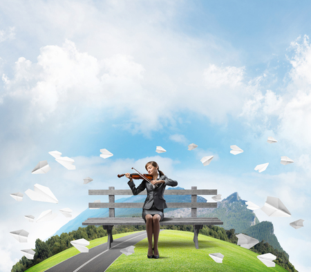 Young woman with violin sitting on wooden bench. Attractive businesslady playing violin. Mountains panorama with round horizon and flying paper planes. Musician practicing and performing outdoors.の写真素材