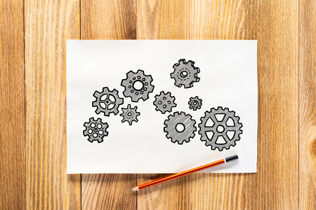 Rotating gears pencil hand drawn. Mechanical technology and machine engineering symbol. Engine mechanism with cogwheels sketch on wooden surface. Workplace with paper and pencil on wooden desk.の写真素材