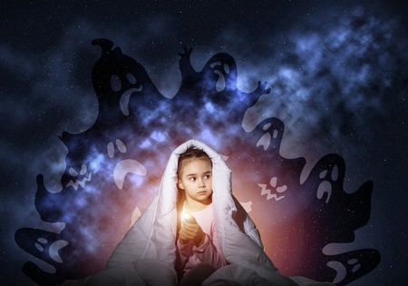 Scared girl with flashlight hiding under blanket from imaginary ghosts. Kid sitting in bed on night sky background. Covered child in pajamas not sleep at night. Mysterious phantoms in darkness.の写真素材