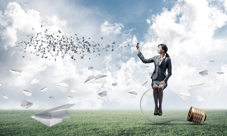 Young woman with smartphone sending text message. Smiling girl in business suit sitting on glowing lamp. Pretty businesswoman using phone on green field with flying paper planes. Mobile communicationの写真素材