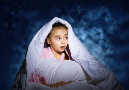 Surprised girl with pillow hiding under blanket. Amazed kid lying in his bed at home. Fear of the dark. Covered kid can not sleep at night. Portrait of girl in pajamas on background of deep blue skyの写真素材
