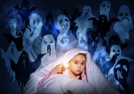 Scared girl with flashlight hiding under blanket from imaginary phantoms. Frightened kid sitting in...