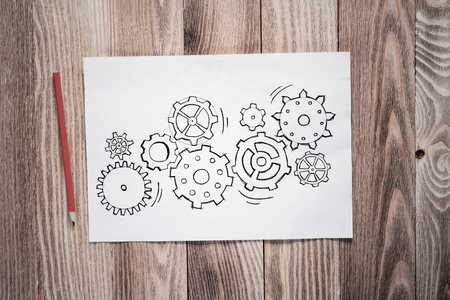 Cogwheels hand drawn illustration on paper sheet. Industry mechanism with cogwheels sketch on wooden surface. Sheet of paper and pencil lying on wooden desk. Textured natural wooden backgroundの写真素材