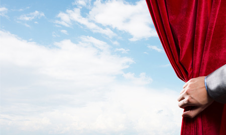 Human hand opens red velvet curtain on blue sky backgroundの写真素材