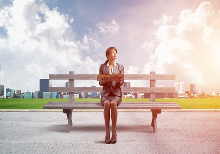 Young woman holding open book and sitting on wooden bench outdoors. Beautiful girl in business suit looking up on background modern cityline panorama in sunny day. Business education and growth.の写真素材