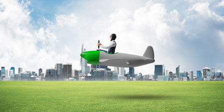 Businessman in aviator hat and goggles driving propeller plane. Downtown with high buildings. Man in airplane flying low above ground. Modern megalopolis panorama with green grass and cloudy skyの写真素材