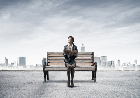 Young woman holding open book and sitting on wooden bench outdoors. Beautiful girl in business suit looking up on background modern cityline panorama with cloudy sky. Business education and growth.の写真素材