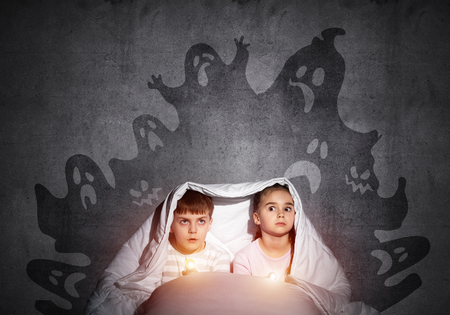 Scared girl and boy with flashlights hiding under blanket from imaginary phantoms. Frightened kids sitting in bed on background grey wall. Children in pajamas and boo ghosts silhouettes above themの写真素材