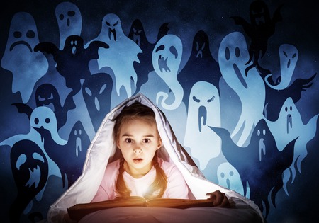 Scared kid with reading book under blanket. Afraid girl lying in bed at home. Child reading scary stories. Girl in pajamas and imaginary ghosts back on night sky. Bright light shining from open book.の写真素材