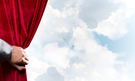 Human hand opens red velvet curtain on blue sky backgroundの写真素材