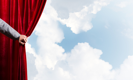 Human hand opens red velvet curtain on blue sky backgroundの写真素材