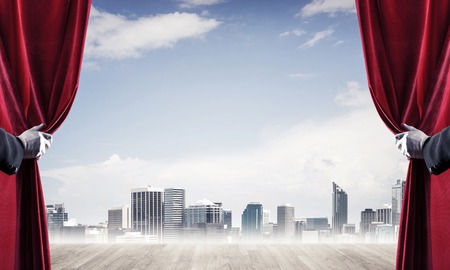 Human hand opens red velvet curtain to modern cityscape and cloudy skyの写真素材