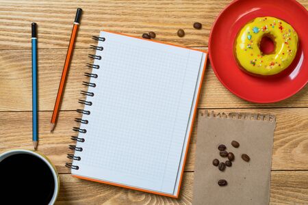 Opened notepad and cup of coffee with donut on wooden tableの写真素材