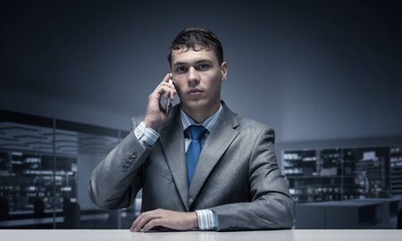 Young man talking on phone and looking at camera. Businessman sitting at desk in office interior....