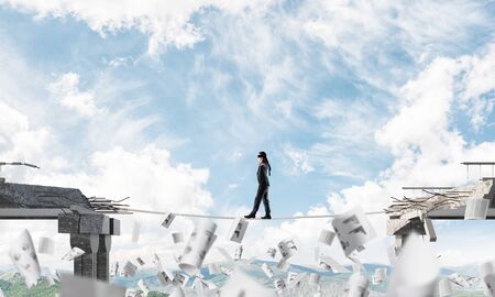 Businessman walking blindfolded on rope among flying papers and above huge gap in bridge as symbol of hidden threats and risks. Nature view on background. 3D rendering.の写真素材