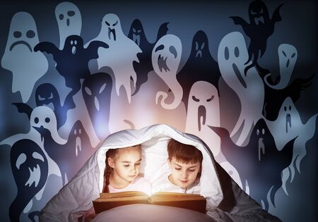 Enthusiastic little girl and boy reading book in bed after bedtime. Kids in pajamas with flashlight hiding under blanket together. Fearful children and imaginary monsters on deep blue background.の写真素材