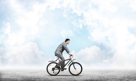 Businessman commuting to work by bike. Man in business suit riding bicycle on road. Handsome cyclist on background of blue sky. Nature landscape with copy space. Eco-friendly transport and active lifeの写真素材