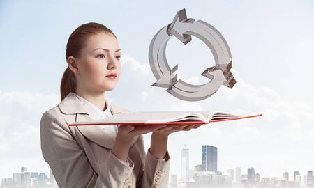 Woman looks at transparent recycle 3d symbol above opened notebook. Renewable energy source concept. Elegant girl with book on background of cityscape and cloudy blue sky.の写真素材